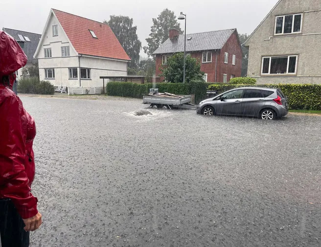 Kvinde i rød regnjakke betragter oversvømmet villavej i voldsomt regnvejr.